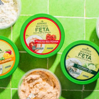 A top-down view of six containers of feta-based spreads arranged on bright green tiled surfaces. Each container has a green lid with varied label colors indicating different flavors, surrounded by the visible textures of the creamy spreads inside.