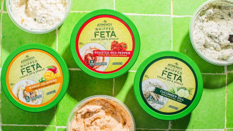 A top-down view of six containers of feta-based spreads arranged on bright green tiled surfaces. Each container has a green lid with varied label colors indicating different flavors, surrounded by the visible textures of the creamy spreads inside.