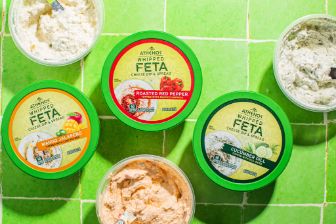 A top-down view of six containers of feta-based spreads arranged on bright green tiled surfaces. Each container has a green lid with varied label colors indicating different flavors, surrounded by the visible textures of the creamy spreads inside.