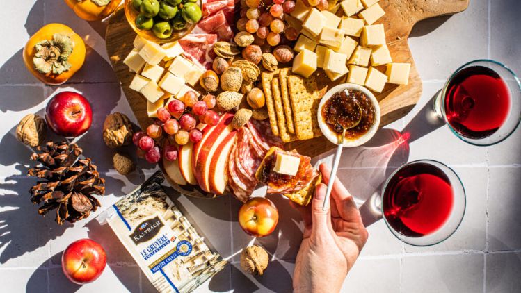 A large wooden charcuterie board arranged with cubes of cheese, assorted crackers, sliced apples, grapes, olives, cured meats, mixed nuts, and a small bowl of fig jam. A hand is spreading jam on a cracker topped with cheese. Surrounding the board on the tiled surface are apples, persimmons, walnuts, pinecones, and a package of crackers. Two glasses filled with a deep red beverage sit nearby, completing the autumn-inspired spread.