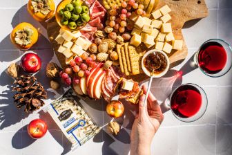A large wooden charcuterie board arranged with cubes of cheese, assorted crackers, sliced apples, grapes, olives, cured meats, mixed nuts, and a small bowl of fig jam. A hand is spreading jam on a cracker topped with cheese. Surrounding the board on the tiled surface are apples, persimmons, walnuts, pinecones, and a package of crackers. Two glasses filled with a deep red beverage sit nearby, completing the autumn-inspired spread.