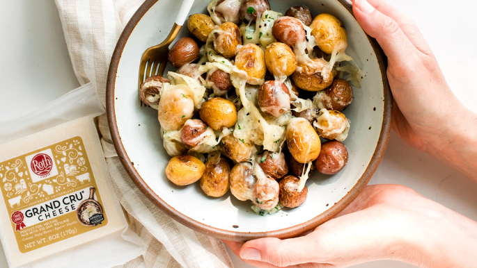 A bowl filled with roasted baby potatoes coated in melted cheese sits on a white surface. A utensil rests inside the bowl, and two hands hold the bowl from each side. To the left, a folded striped kitchen towel lies next to a package of Roth Grand Cru cheese. The bright, clean setting highlights the golden potatoes and creamy melted cheese.
