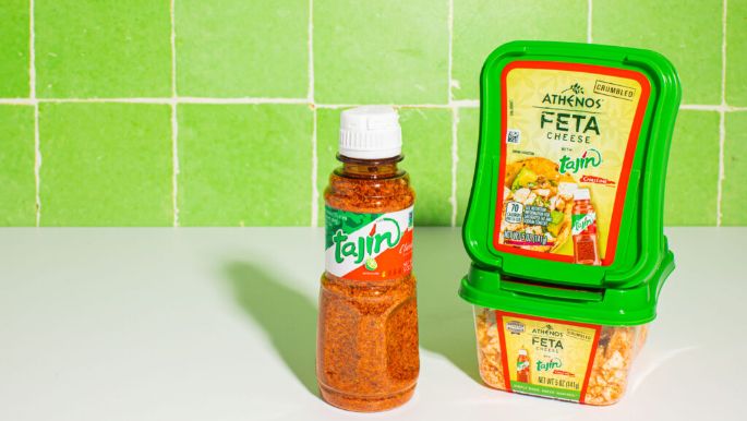 A container of Athenos Feta Cheese with Tajín seasoning is displayed next to a bottle of Tajín seasoning on a white surface, with bright green tiled walls in the background. The feta container has a green lid and a colorful label showing serving suggestions, while the Tajín bottle contains reddish seasoning with a white cap. Both products are well-lit against the clean, minimal backdrop.