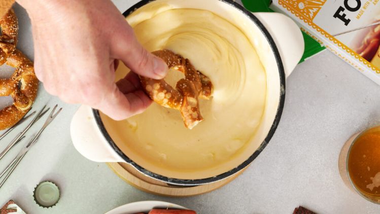 A game‑day snack spread featuring soft pretzels, mini sausages, crackers, a bowl of dip, beer fondue packaging, beverage glasses, and bottle caps arranged on a light tabletop with football‑themed accents.
