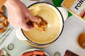 A game‑day snack spread featuring soft pretzels, mini sausages, crackers, a bowl of dip, beer fondue packaging, beverage glasses, and bottle caps arranged on a light tabletop with football‑themed accents.