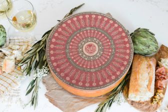 A large wheel of Roth cheese with an ornate red and beige patterned label sits on a rustic white tabletop. Surrounding the cheese wheel are artichokes, a crusty loaf of bread, thin slices of cured meat, fresh herbs, small white flowers, and two glasses filled with a pale beverage. Bits of broken cheese and a striped cloth add to the styled, artisanal presentation.