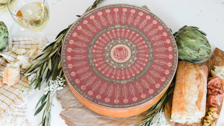 A large wheel of Roth cheese with an ornate red and beige patterned label sits on a rustic white tabletop. Surrounding the cheese wheel are artichokes, a crusty loaf of bread, thin slices of cured meat, fresh herbs, small white flowers, and two glasses filled with a pale beverage. Bits of broken cheese and a striped cloth add to the styled, artisanal presentation.