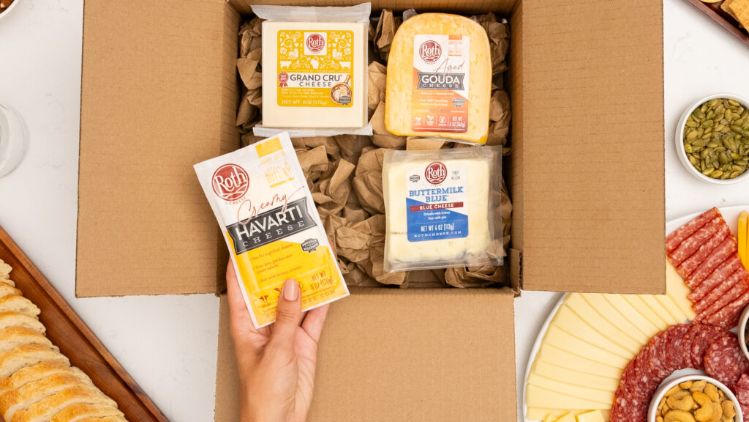 A cardboard box filled with assorted Roth cheese varieties sits open on a white tabletop. Inside the box are three different packaged cheeses surrounded by brown packing paper. A hand is placing an additional Roth Havarti cheese block into the box. Around the box are various foods including sliced bread, nuts, crackers, cured meats, cheese slices, pickles, and olives, arranged in small bowls and on serving platters.