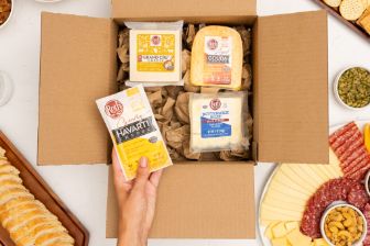 A cardboard box filled with assorted Roth cheese varieties sits open on a white tabletop. Inside the box are three different packaged cheeses surrounded by brown packing paper. A hand is placing an additional Roth Havarti cheese block into the box. Around the box are various foods including sliced bread, nuts, crackers, cured meats, cheese slices, pickles, and olives, arranged in small bowls and on serving platters.