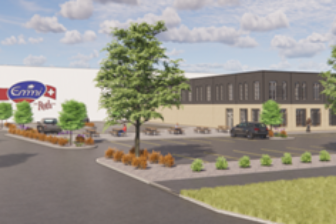 A wide view of an Emmi Roth facility surrounded by landscaped greenery, parking areas, and trees, featuring a prominent Emmi Roth sign near the entrance under a bright, partly cloudy sky.