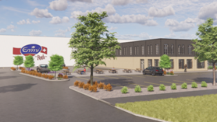 A wide view of an Emmi Roth facility surrounded by landscaped greenery, parking areas, and trees, featuring a prominent Emmi Roth sign near the entrance under a bright, partly cloudy sky.