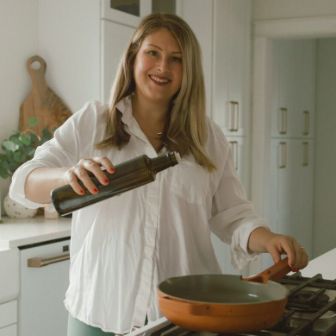 A person in a bright, modern kitchen pouring oil from a dark glass bottle into a skillet on a stovetop, with countertops, cabinets, and cooking tools visible in the background.