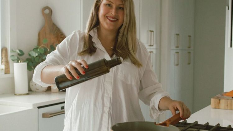 A person in a bright, modern kitchen pouring oil from a dark glass bottle into a skillet on a stovetop, with countertops, cabinets, and cooking tools visible in the background.