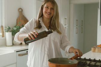 A person in a bright, modern kitchen pouring oil from a dark glass bottle into a skillet on a stovetop, with countertops, cabinets, and cooking tools visible in the background.