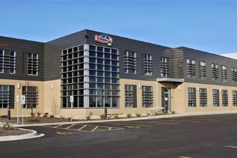 Emmi Roth Stoughton Building. A modern industrial building with a beige lower façade and dark gray upper structure, featuring large grid-style windows on both floors. The building sits beside a wide, empty asphalt parking lot with marked parking spaces, landscaped areas, and a small number of young trees. A sign with a red-and-blue logo is mounted near the roofline of the central section.