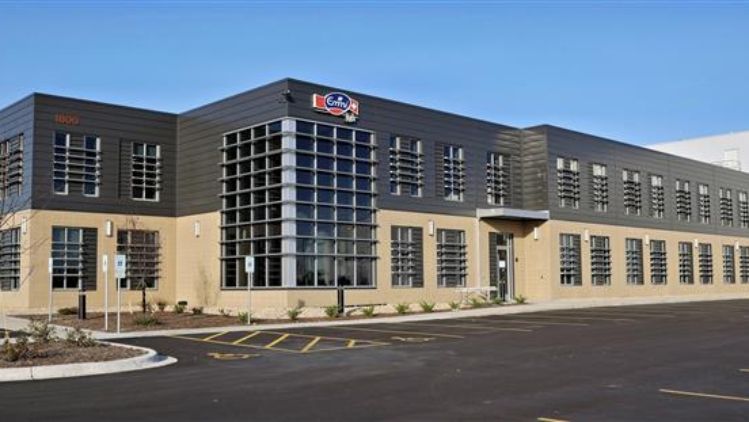 Emmi Roth Stoughton Building. A modern industrial building with a beige lower façade and dark gray upper structure, featuring large grid-style windows on both floors. The building sits beside a wide, empty asphalt parking lot with marked parking spaces, landscaped areas, and a small number of young trees. A sign with a red-and-blue logo is mounted near the roofline of the central section.