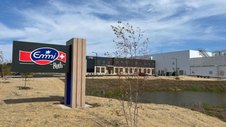 A modern Emmi Roth facility featuring a prominent Emmi Roth sign in the foreground, set against industrial buildings, open landscaping, and a clear blue sky.