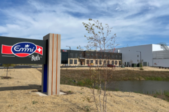 A modern Emmi Roth facility featuring a prominent Emmi Roth sign in the foreground, set against industrial buildings, open landscaping, and a clear blue sky.