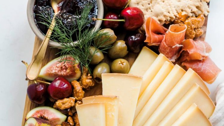 A beautifully arranged cheeseboard featuring neatly sliced cheese alongside figs, cherries, olives, cured meats, nuts, crisp crackers, and a bowl of dark fruit jam, all presented on a wooden serving board.