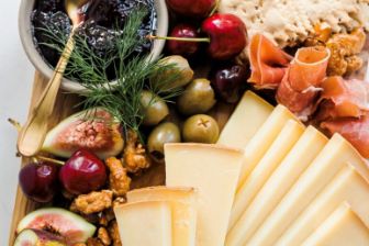 A beautifully arranged cheeseboard featuring neatly sliced cheese alongside figs, cherries, olives, cured meats, nuts, crisp crackers, and a bowl of dark fruit jam, all presented on a wooden serving board.
