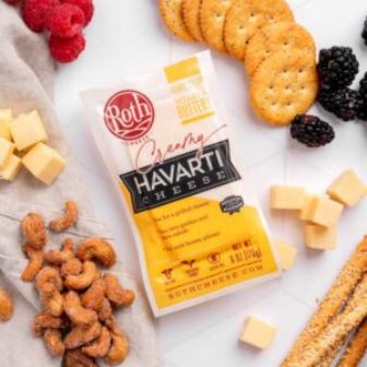 A package of Roth Creamy Havarti cheese placed on a white surface surrounded by crackers, blackberries, raspberries, cheese cubes, seasoned nuts, and crispy breadsticks, creating a bright and inviting snacking arrangement.