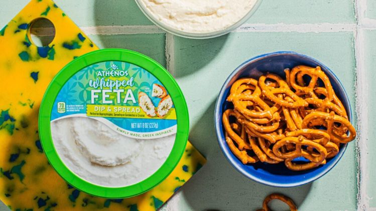 A container of Athenos Whipped Feta Dip &amp; Spread displayed on a vibrant cutting board alongside a bowl of pretzels, with an additional bowl of whipped feta in the background on a tiled surface.
