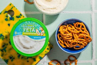 A container of Athenos Whipped Feta Dip &amp; Spread displayed on a vibrant cutting board alongside a bowl of pretzels, with an additional bowl of whipped feta in the background on a tiled surface.