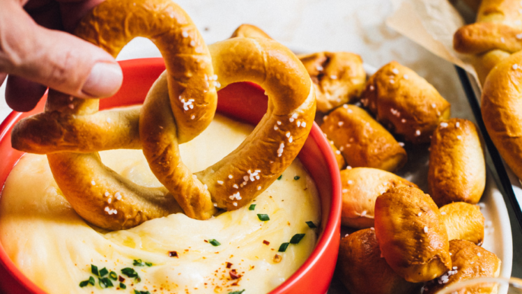 A warm soft pretzel being dipped into a creamy cheese sauce in a red bowl, surrounded by additional pretzels and bite‑sized pretzel pieces on a light surface.