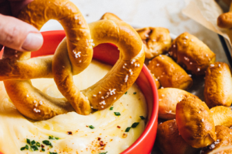 A warm soft pretzel being dipped into a creamy cheese sauce in a red bowl, surrounded by additional pretzels and bite‑sized pretzel pieces on a light surface.