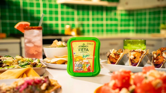 A container of Athenos Feta Cheese with Tajín seasoning is placed at the center of a brightly lit kitchen scene. Surrounding it are various colorful dishes, including tacos, salads, and bowls of fresh ingredients. A pink beverage with a salt‑rimmed glass sits in the background, along with green tiled walls, utensils, and blurred kitchen items.
