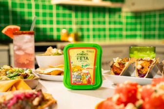 A container of Athenos Feta Cheese with Tajín seasoning is placed at the center of a brightly lit kitchen scene. Surrounding it are various colorful dishes, including tacos, salads, and bowls of fresh ingredients. A pink beverage with a salt‑rimmed glass sits in the background, along with green tiled walls, utensils, and blurred kitchen items.