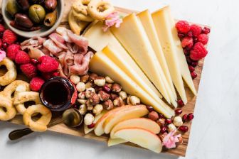 A wooden serving board arranged with slices of semi-hard cheese, fresh raspberries, apple slices, olives, cured meats, round bread snacks, mixed nuts, and a small bowl of dark dipping sauce.