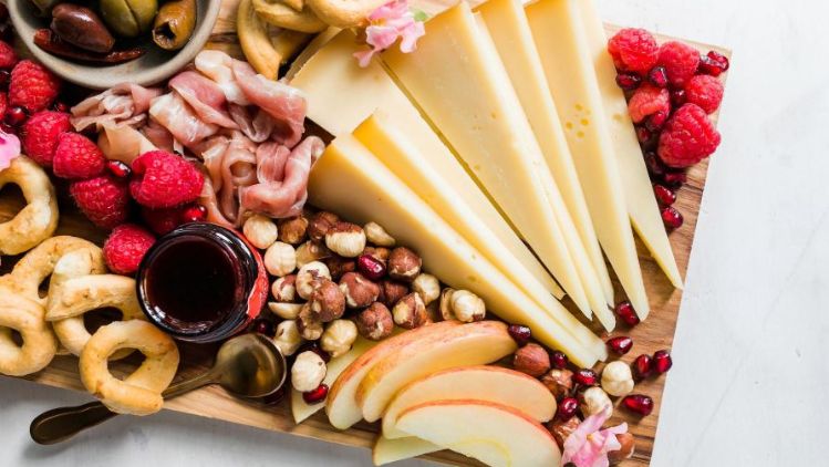 A wooden serving board arranged with slices of semi-hard cheese, fresh raspberries, apple slices, olives, cured meats, round bread snacks, mixed nuts, and a small bowl of dark dipping sauce.