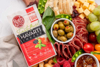 A colorful charcuterie board featuring Roth Tomato Basil Havarti cheese alongside sliced bread, olives, cured meats, fresh fruits, vegetables, crackers, and a fig spread, arranged on a wooden serving board.