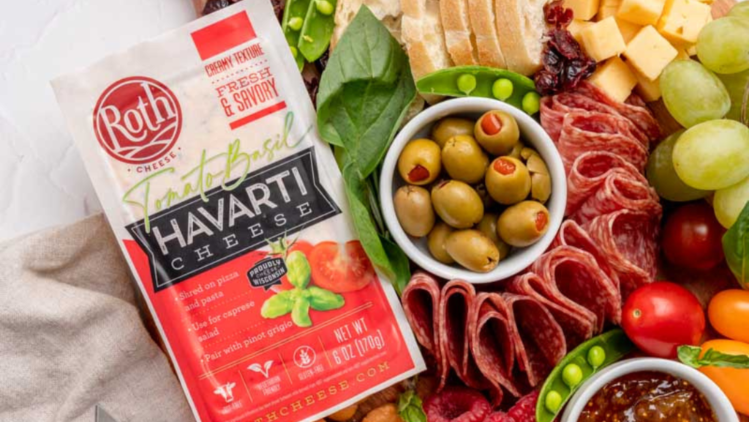 A colorful charcuterie board featuring Roth Tomato Basil Havarti cheese alongside sliced bread, olives, cured meats, fresh fruits, vegetables, crackers, and a fig spread, arranged on a wooden serving board.